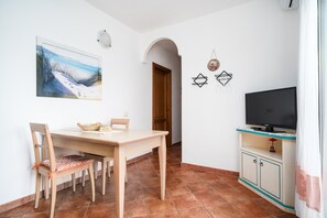 A dining area with a wooden table and chairs, a painting on the wall, a television on a cabinet, and a door leading to another room.