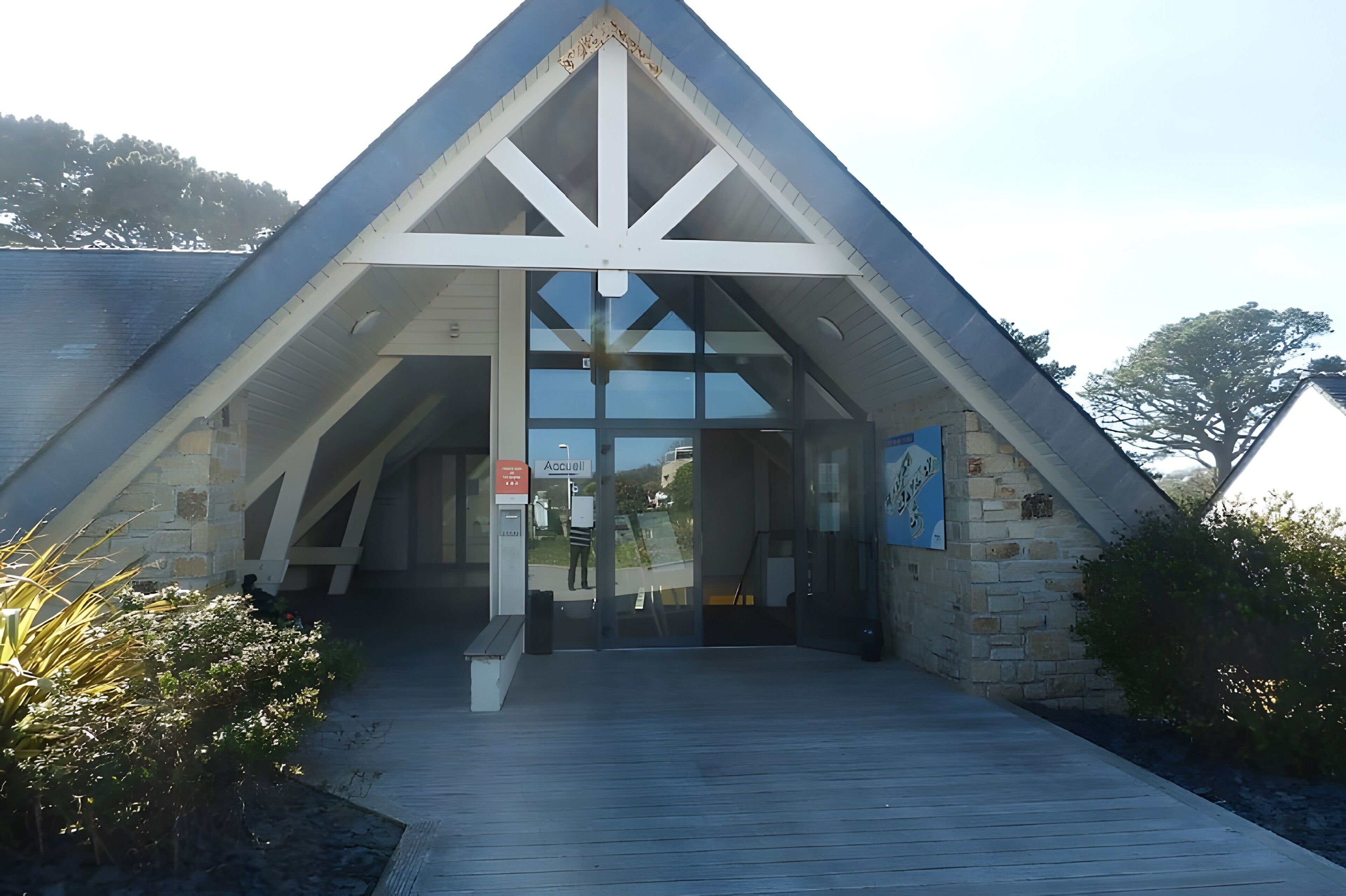 A building with a triangular roof, a glass entrance, and a wooden walkway leading to it.