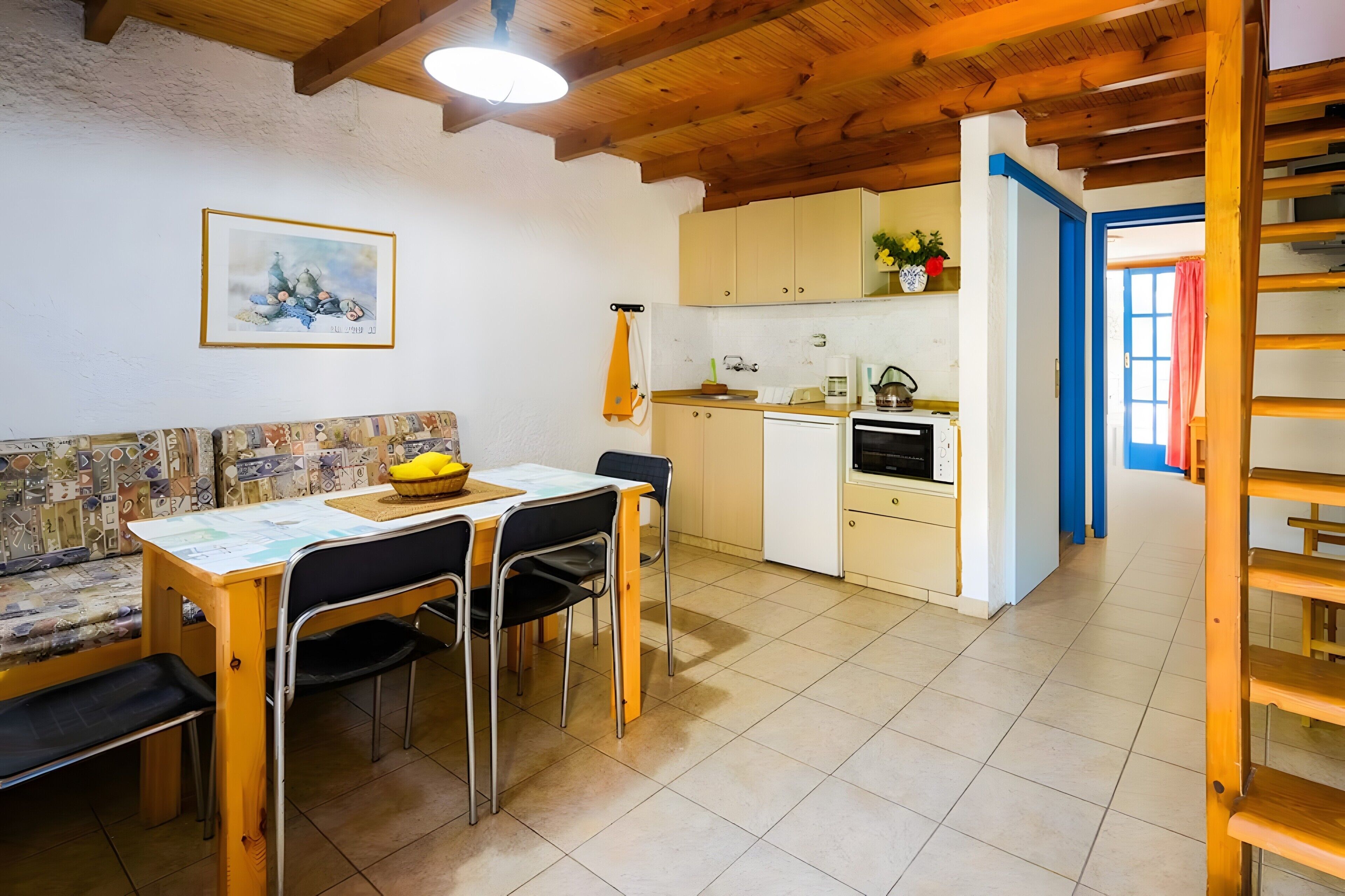Il y a un coin repas avec une table, des chaises et un banc. On y trouve également une cuisine équipée de placards et d’une cuisinière. Un tableau est accroché au mur, et une porte donne accès à une autre pièce.