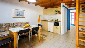 Il y a un coin repas avec une table, des chaises et un banc. On y trouve également une cuisine équipée de placards et d’une cuisinière. Un tableau est accroché au mur, et une porte donne accès à une autre pièce.
