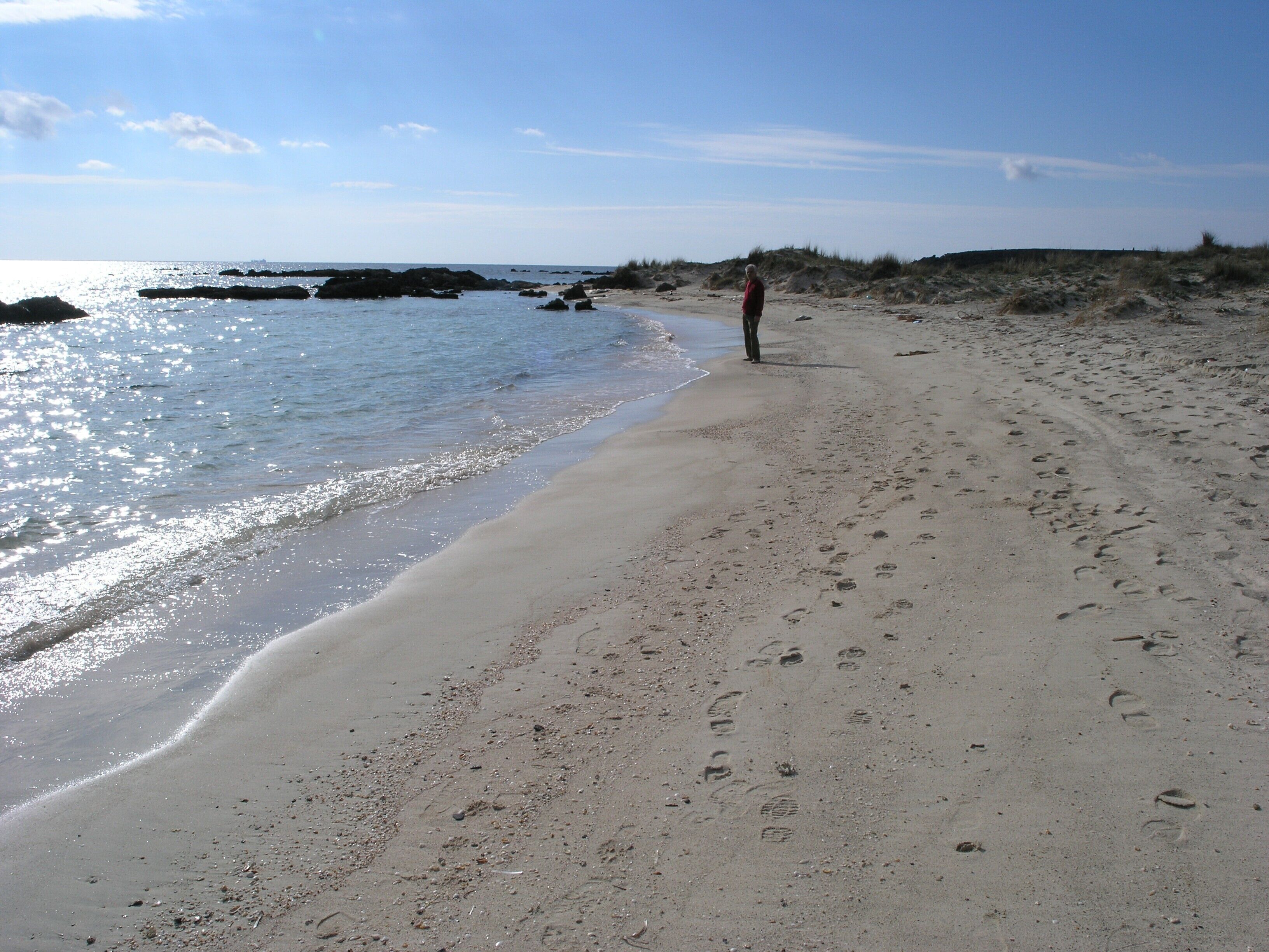 Une plage où se trouve une personne debout, avec un sol sablonneux et des affleurements rocheux dans l’eau.