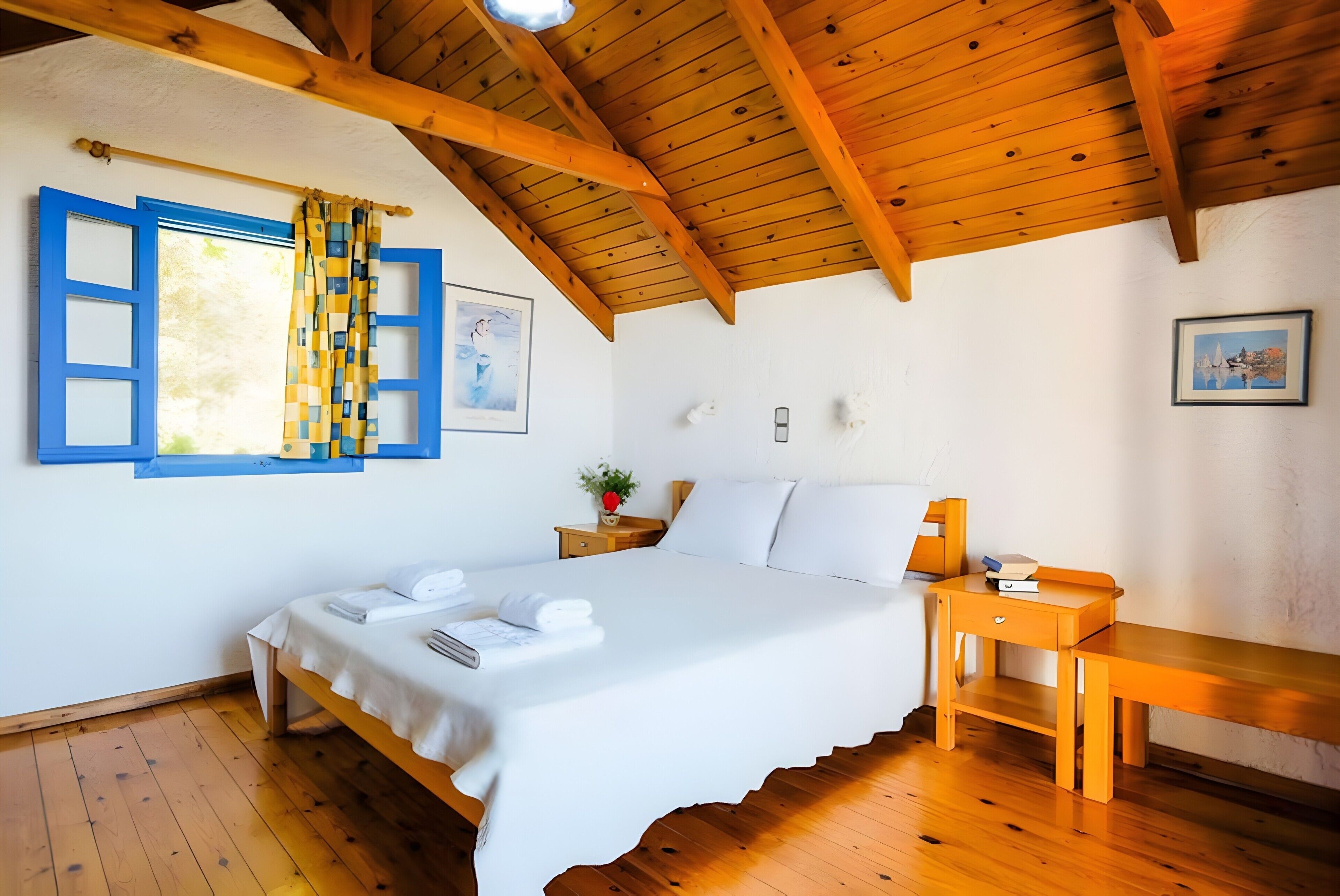 A bedroom with a wooden ceiling, a bed with white linens, a wooden nightstand, and a window with blue shutters.