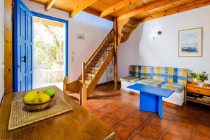 Una habitación con escalera de madera, puerta azul, suelo de baldosas, un banco con cojín de cuadros y una mesa con cesta de fruta.