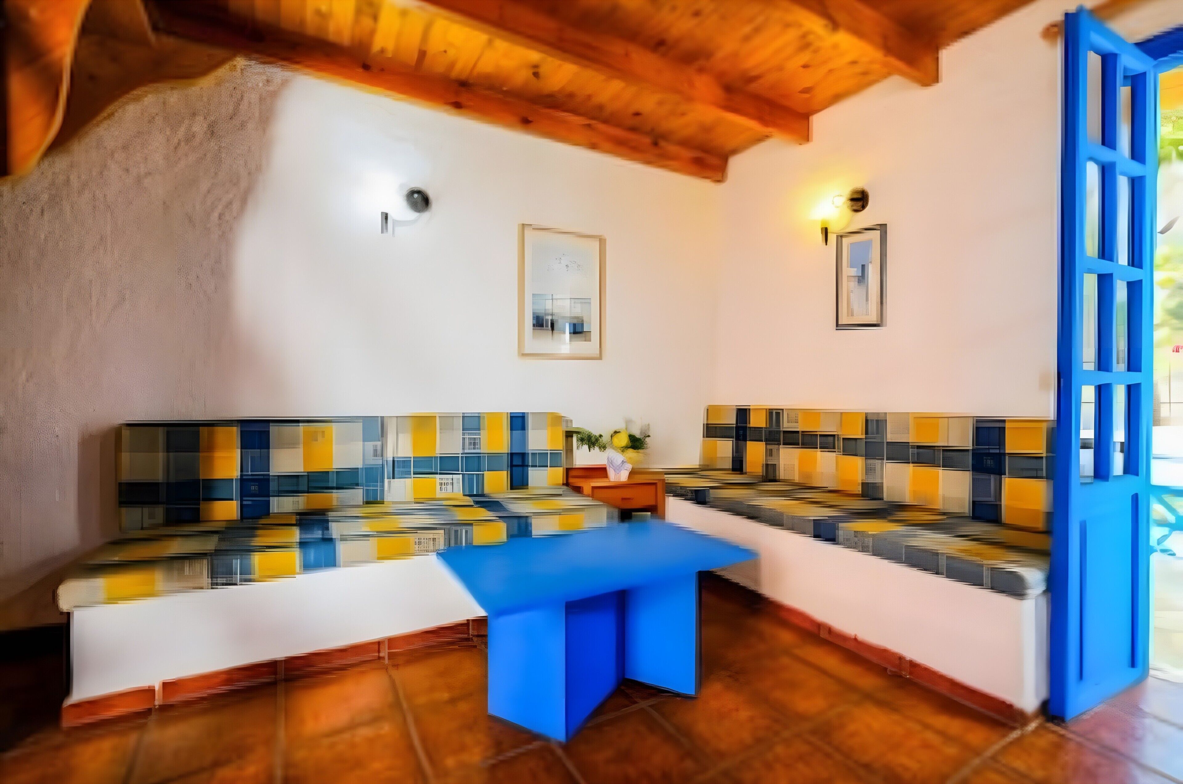 A room with a wooden ceiling, a blue door, a white wall, and a sofa with a yellow and blue patterned cushion.