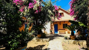 A courtyard with a stone pathway leading to a building with a blue door, surrounded by potted plants and flowering trees. - Cottage 'Olive Tree   4' with Sea View, Private Terrace and Wi-Fi (Paleochora-Crete)