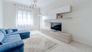 A modern living room with a large flat-screen TV, a blue sofa set, a light wooden TV stand, and a patterned rug. - GuestHost - Comfy APT in Santa Rita District (Torino)