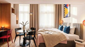 Ein modernes Hotelzimmer mit Bett, Nachttisch, Leselampe sowie einer Essecke mit Stühlen und Tisch.