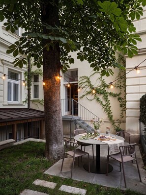 Une table ronde entourée de chaises, installée à l’extérieur, avec un arbre et des guirlandes lumineuses en arrière-plan.
