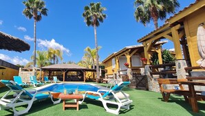 Un espace piscine avec des chaises longues, un parasol en chaume et un bâtiment à la façade en pierre.