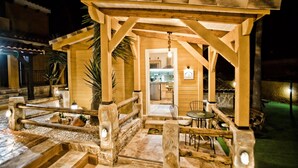 Une pergola en bois abritant un espace repas, des murs en pierre et, au fond, une cuisine ouverte.