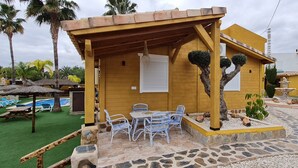 A yellow building with a covered patio, outdoor seating, a swimming pool, and palm trees.
