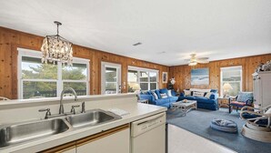 Una cucina moderna con lavello, lavastoviglie e pareti in legno. C'è un'area soggiorno con divani blu e un quadro raffigurante una spiaggia.