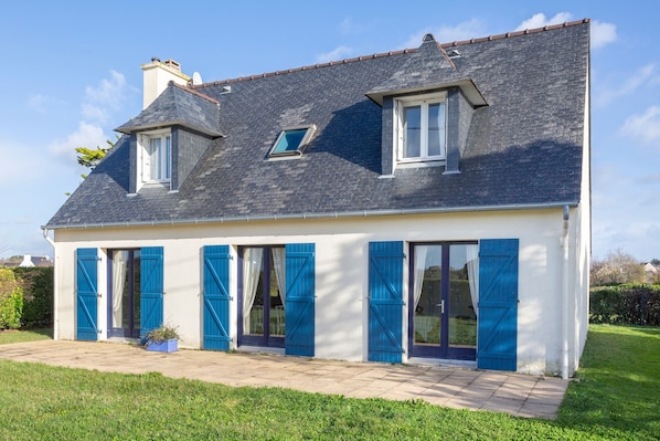 Une maison à deux étages avec un toit en pente, de grandes fenêtres et des volets bleus.