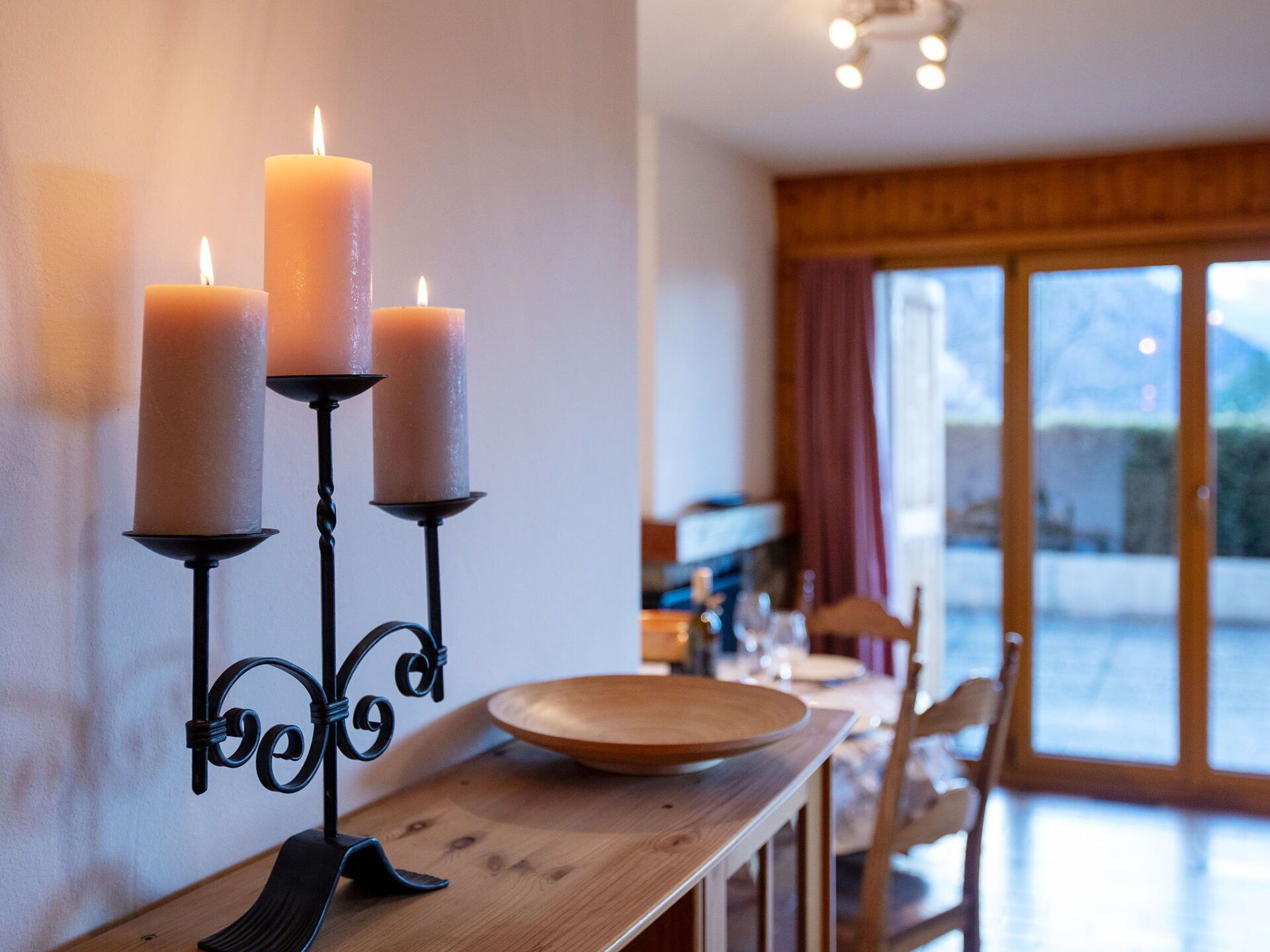 A dining area with a wooden table, three candles on a wall-mounted holder, and a view of the outdoors through large windows.