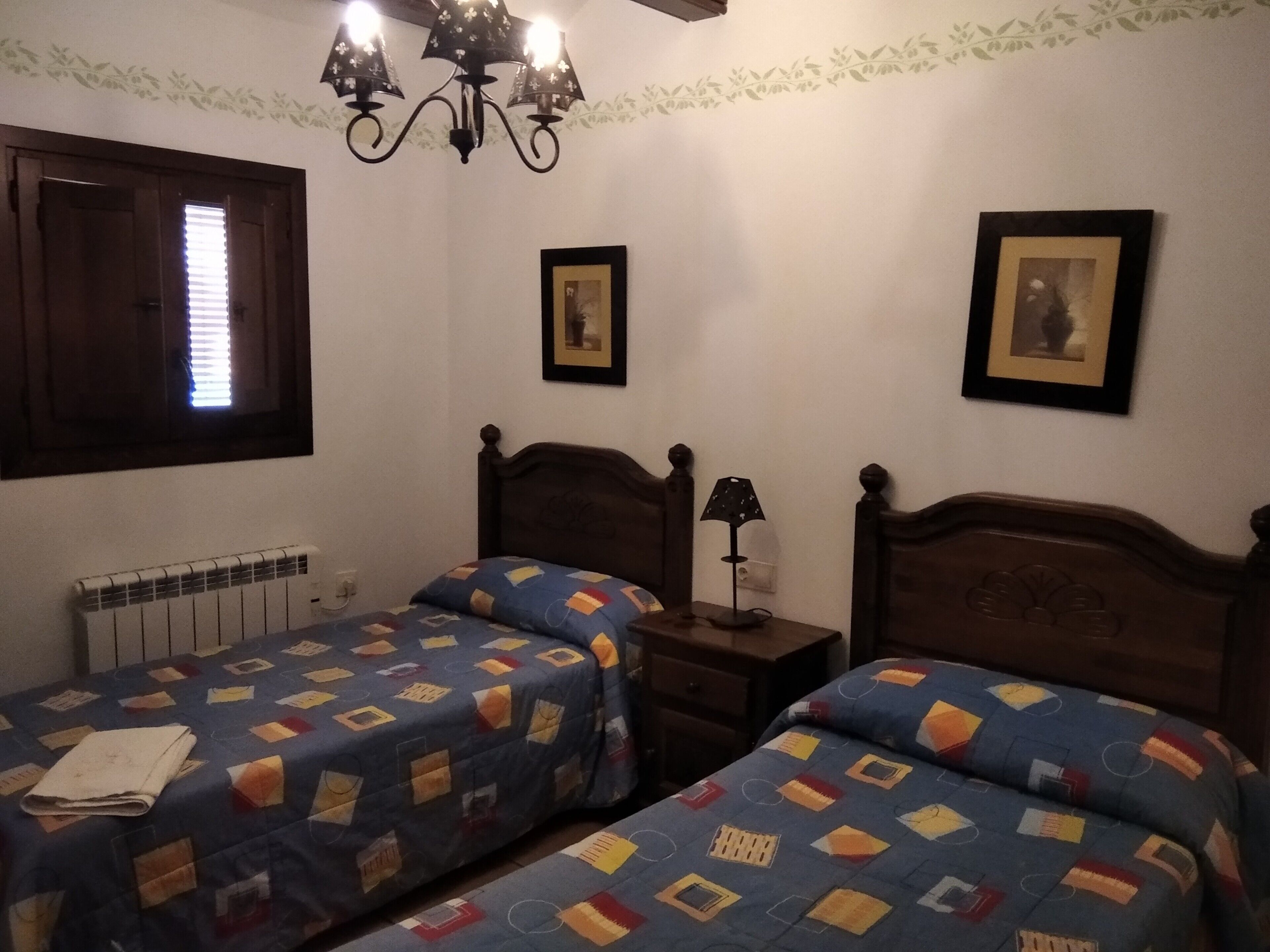 Una camera da letto con due letti, un termosifone, una finestra con persiane e una lampada a parete, oltre a quadri appesi.
