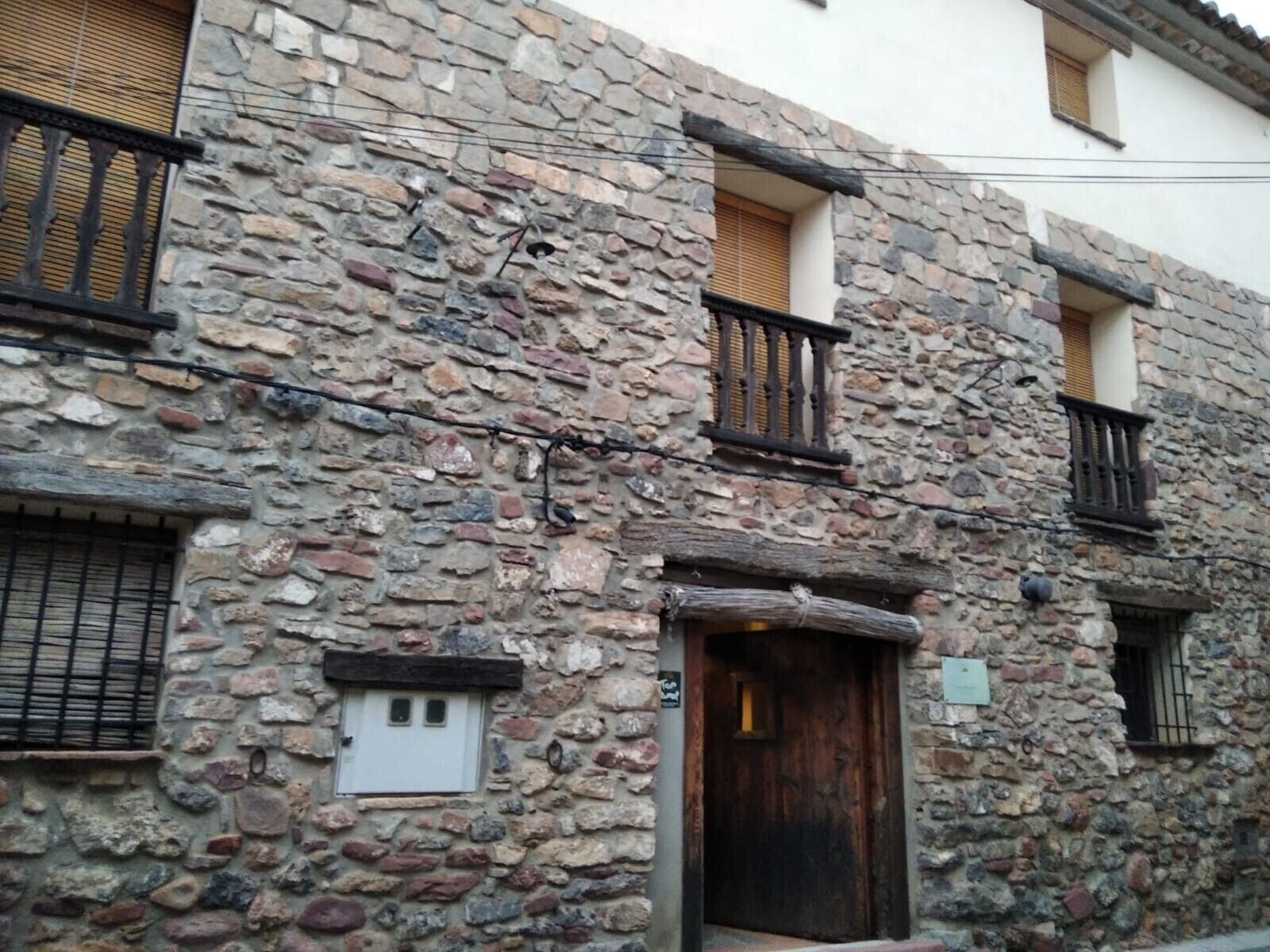 Un edificio de piedra con puerta de madera, una ventana pequeña con persiana y un balcón con barandilla de metal.