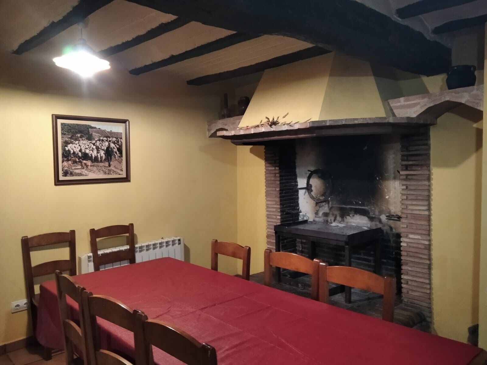 A dining area with a red tablecloth, wooden chairs, a brick fireplace, and a framed picture on the wall.