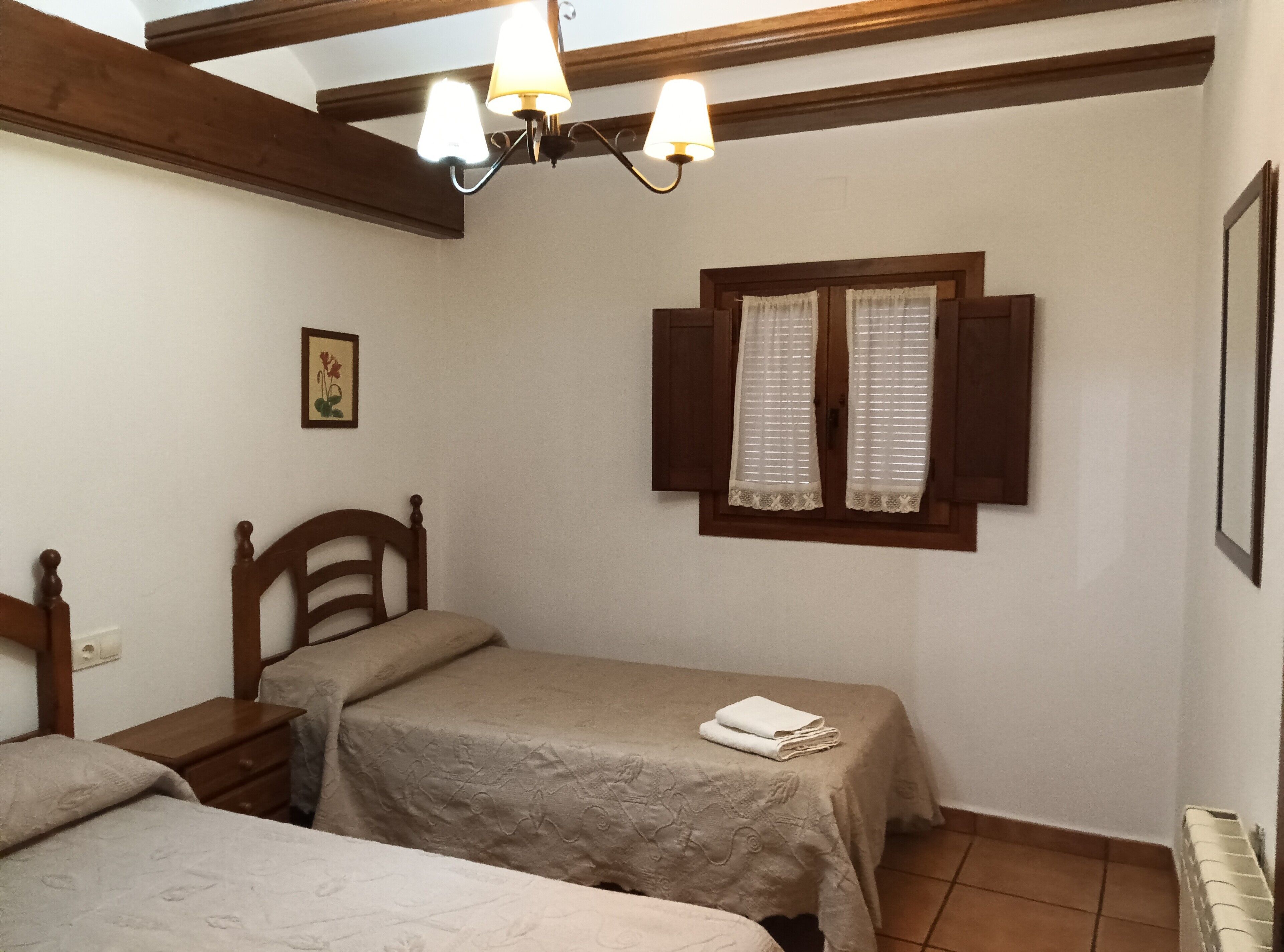 A bedroom with two beds, a wooden headboard, a nightstand, a window with shutters, a wall-mounted light fixture, and a framed picture.