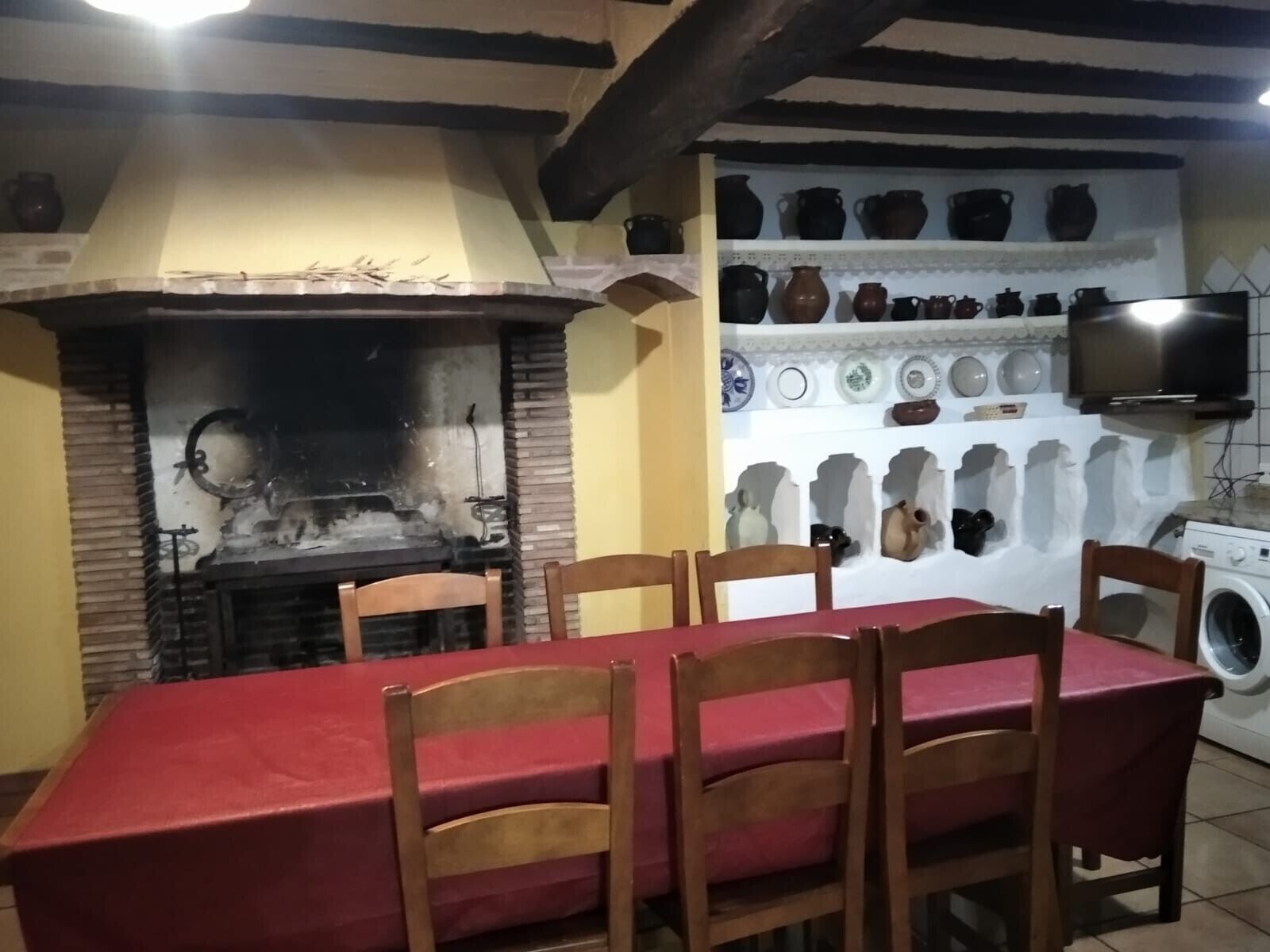 Un coin repas avec une table rouge, des chaises en bois, une cheminée en briques et un mur décoré de poteries.