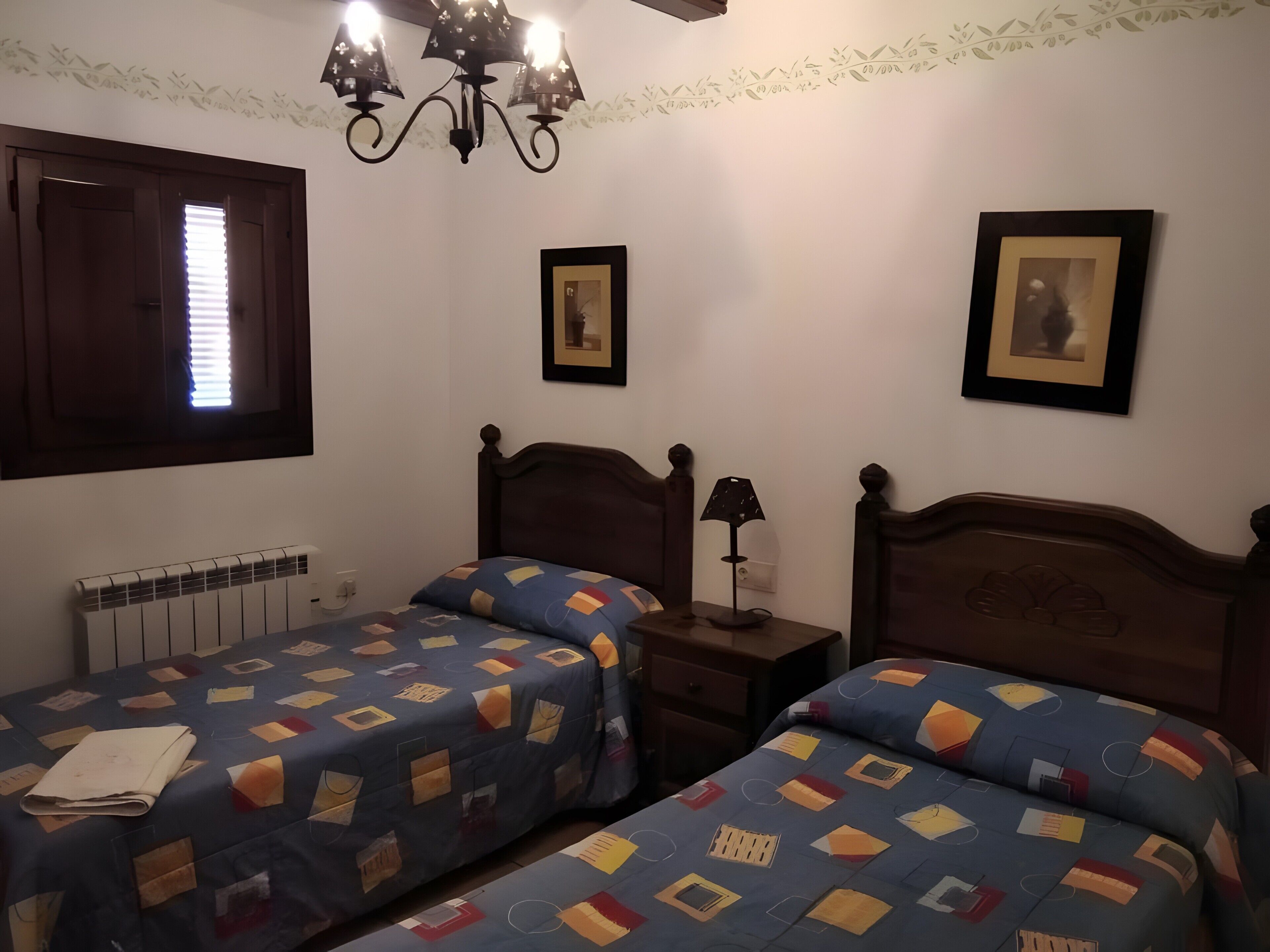 Un dormitorio con dos camas, un radiador, una lámpara fijada a la pared y cuadros enmarcados.