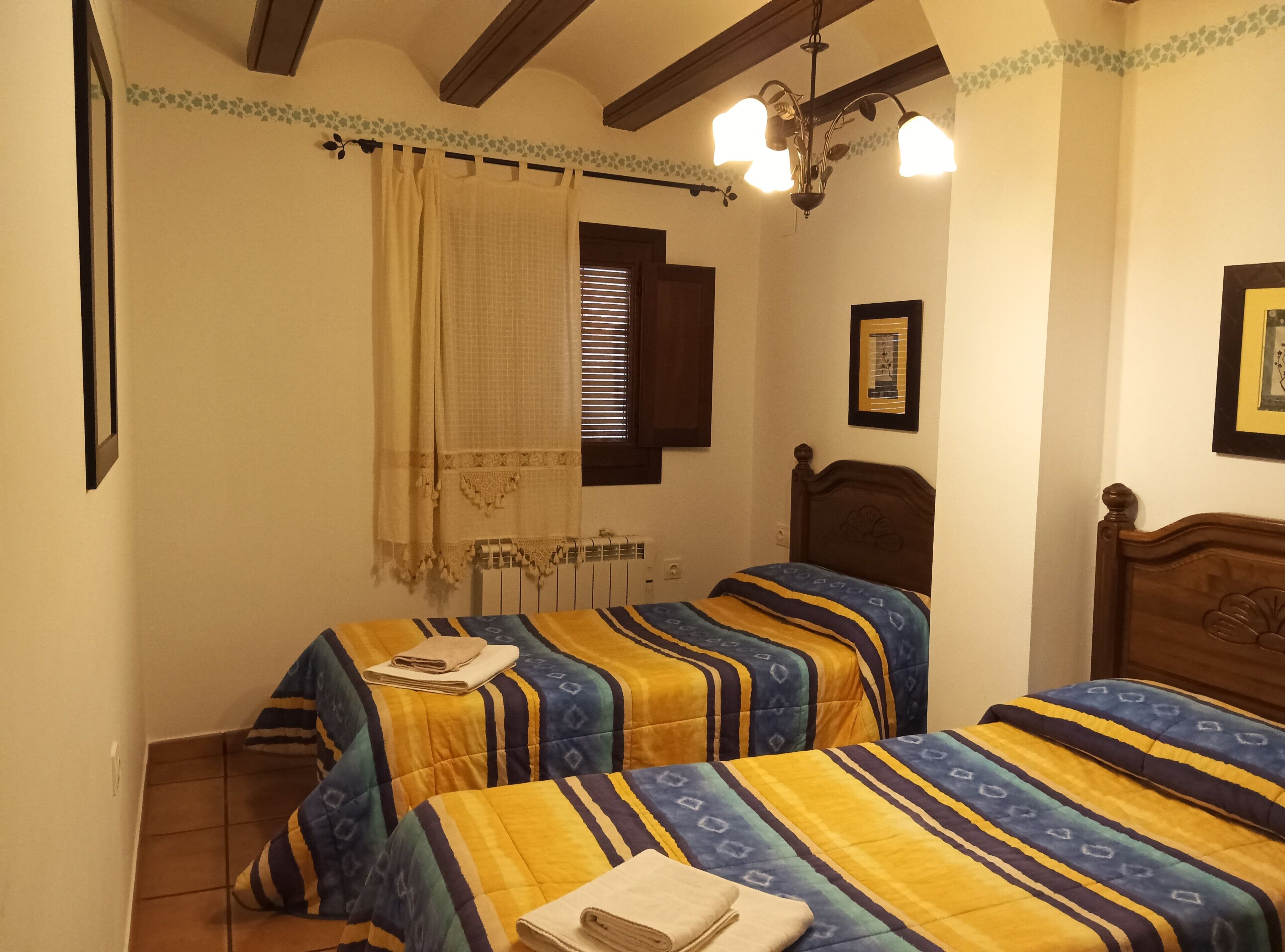 Camera da letto con due letti, un termosifone, una finestra con persiane, un lampadario e un quadro appeso al muro.