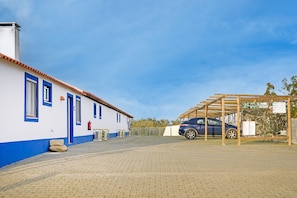 A building with a blue door and windows, a car parked under a wooden structure, and a paved area.