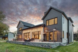 A modern two-story house with a stone foundation, large windows, and a patio with outdoor furniture.
