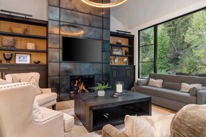 A modern living room with a fireplace, sofas, and a large window overlooking a forest.