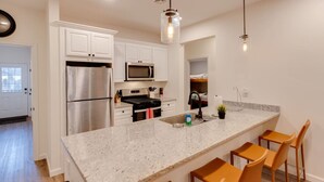 A modern kitchen with white cabinets, stainless steel appliances, a granite countertop, and wooden bar stools. - Luxe Condo-Shuttle & Amenities (Lincoln)