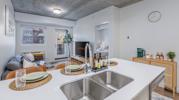 Una cocina moderna con una isla grande, un lavamanos de acero inoxidable y un horno empotrado. Hay un comedor con una mesa para dos, y una sala de estar con un sofá y un televisor.