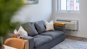 Una sala de estar moderna con un sofá gris, un aire acondicionado blanco y vistas de edificios a través de una ventana.