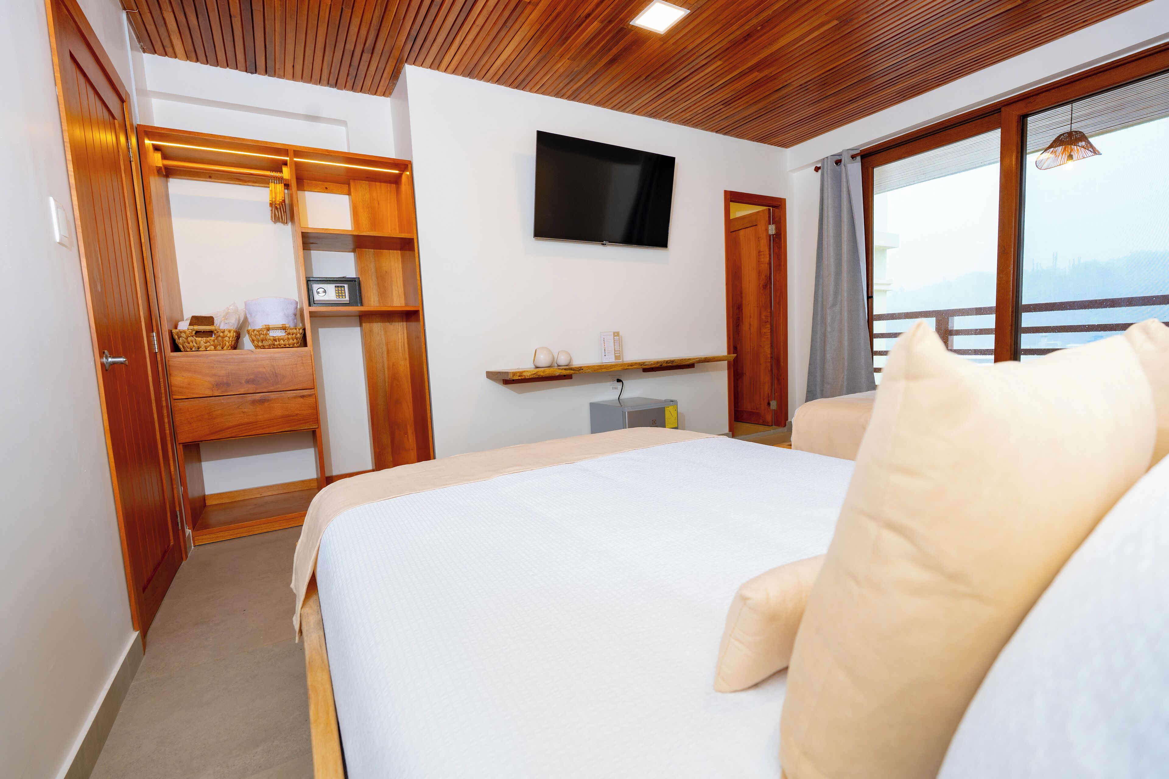 A bedroom with a bed, a TV mounted on the wall, a wooden wardrobe, and a window with a view.