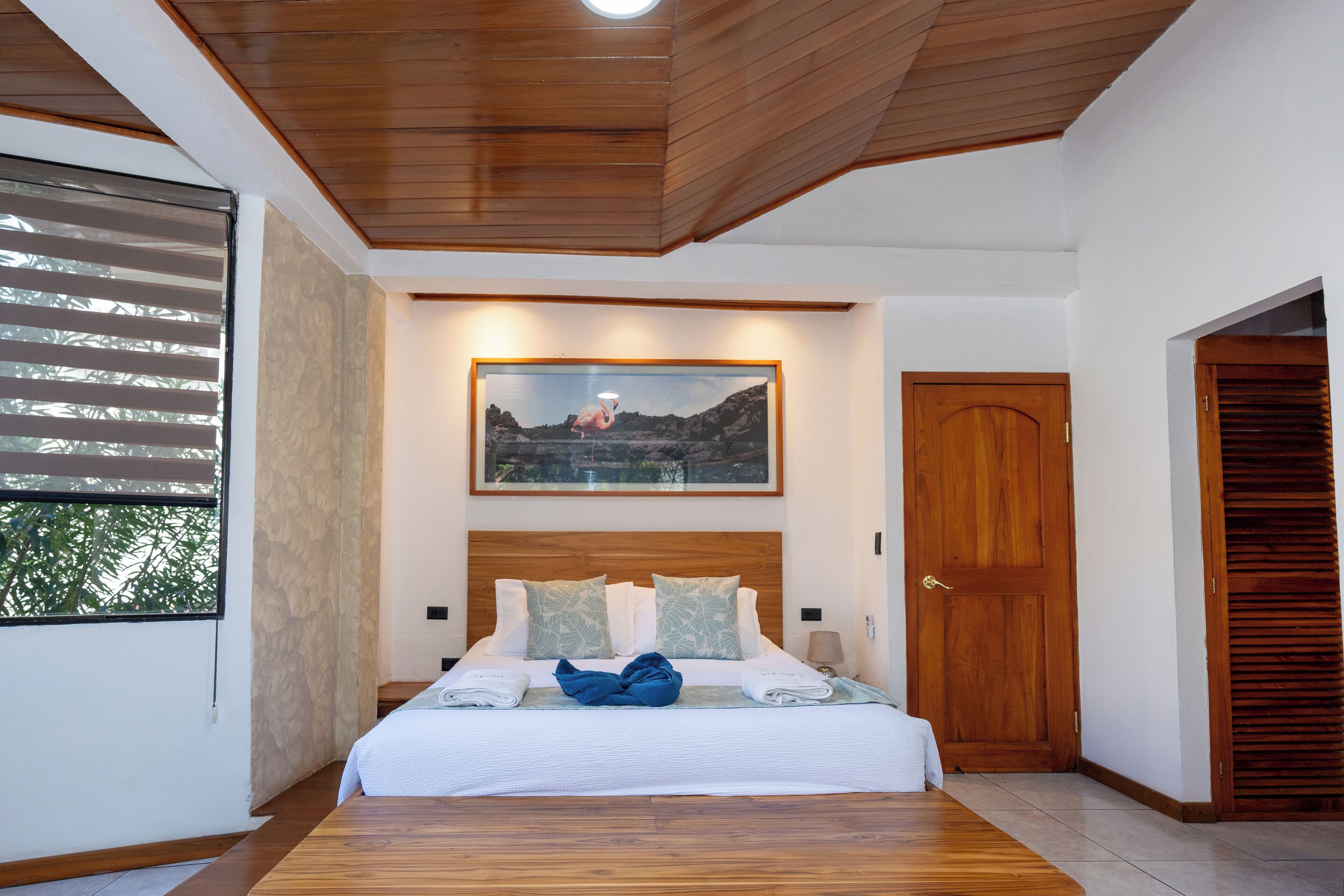 A bedroom with a wooden ceiling, a large bed with a white bedspread, two pillows, and a framed picture above the bed.