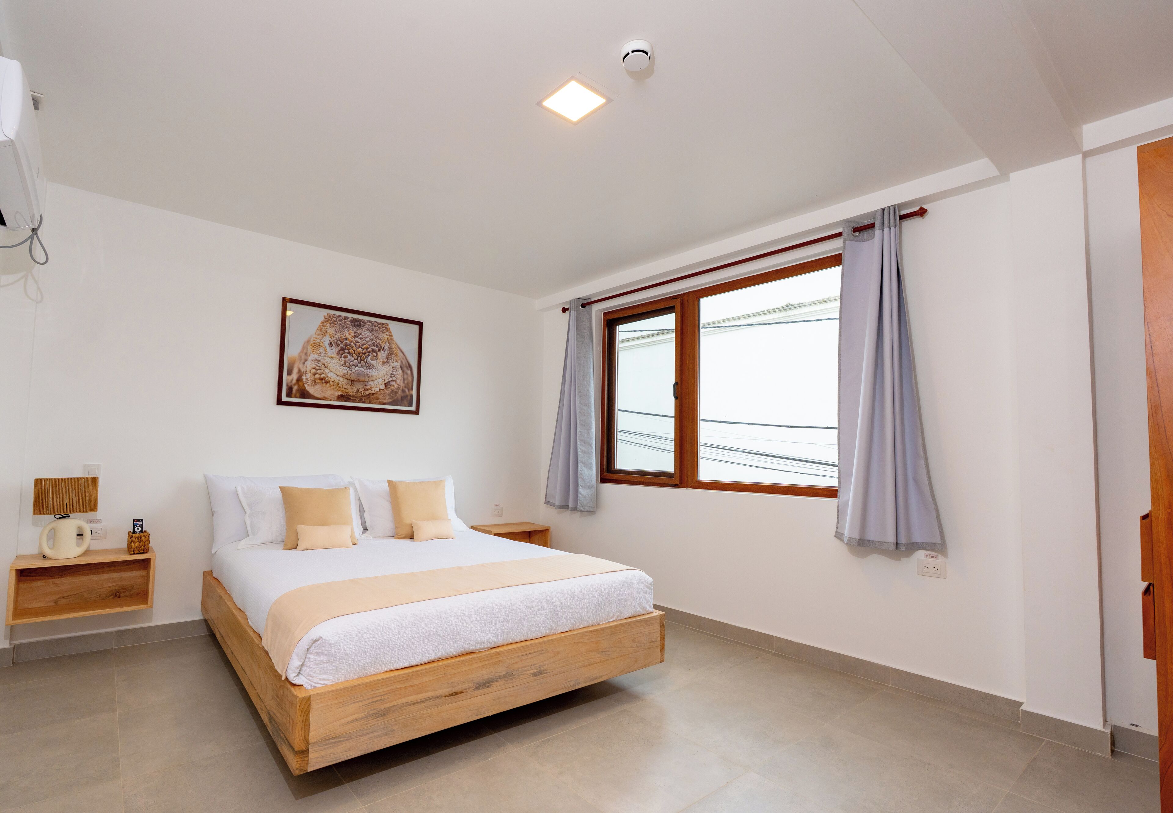 Un dormitorio con una cama de madera, una mesita de noche con lámpara, un cuadro en la pared y una ventana con cortinas.