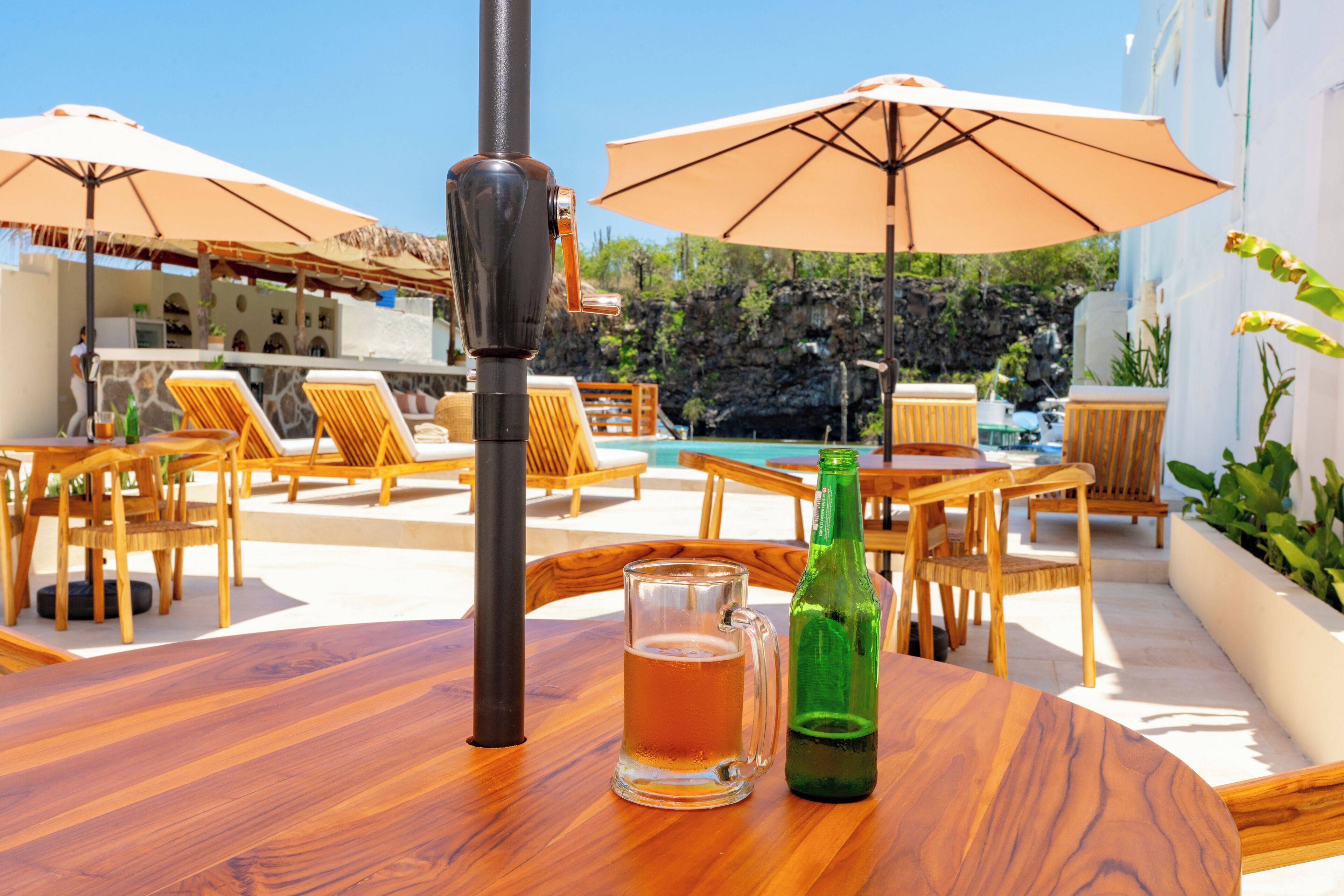Una mesa de madera con una jarra de cerveza y una botella, rodeada de sillones de exterior y sombrillas, con vistas a una piscina y vegetación.