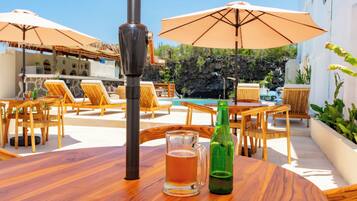 A wooden table with a beer mug and a bottle, surrounded by lounge chairs and umbrellas, overlooking a pool and greenery.