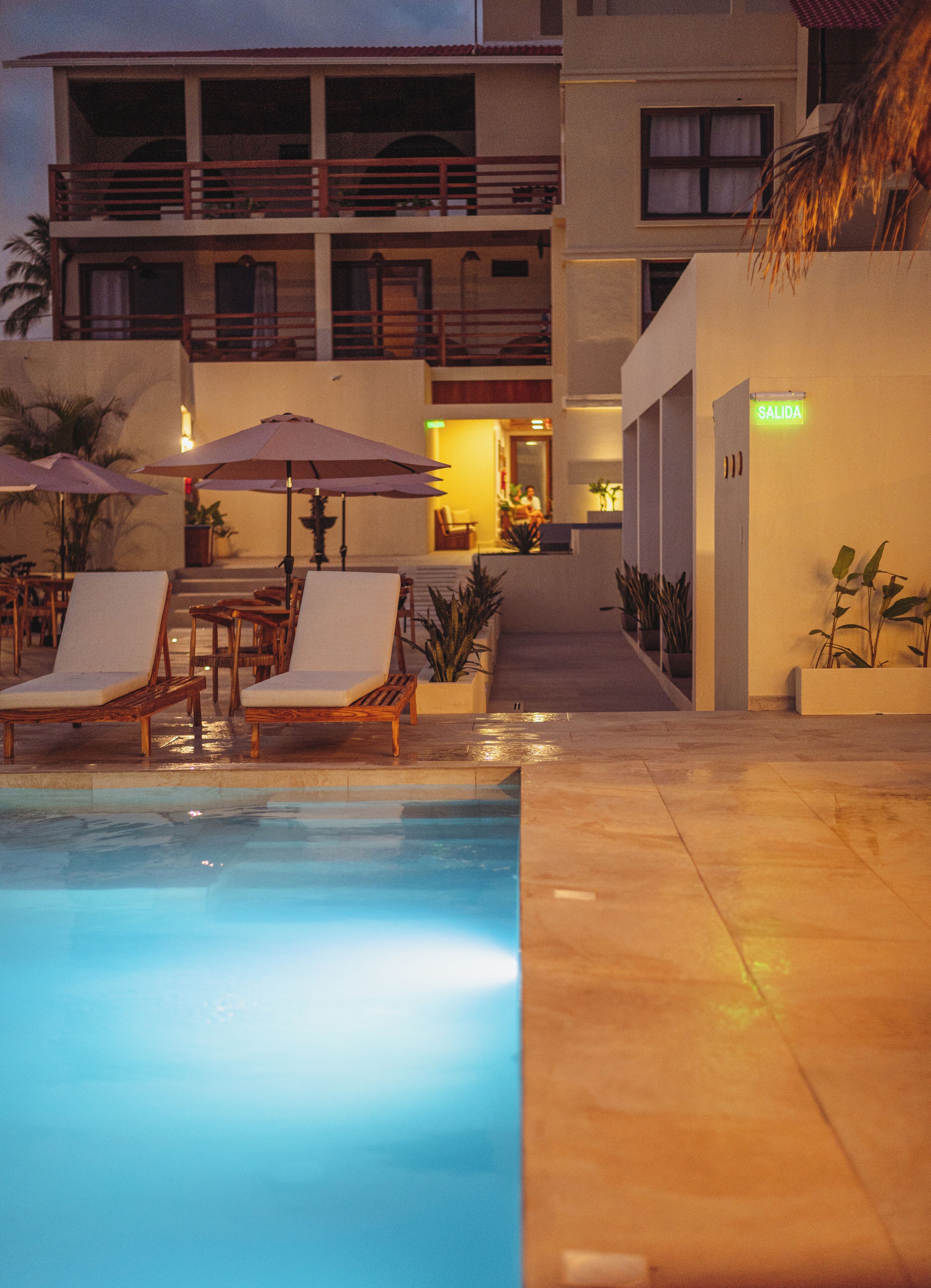 A pool area with lounge chairs and umbrellas, a building with a 'SALIDA' sign, and a staircase leading to an upper level.