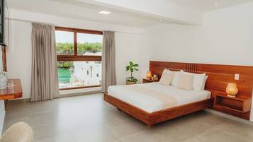 A bedroom with a large bed, a TV, a desk, a chair, a window with curtains, and a view of greenery outside.