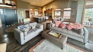 A spacious living area with a large sofa, a smaller sofa, and a coffee table. There is a kitchen area in the background with a dining table and chairs.