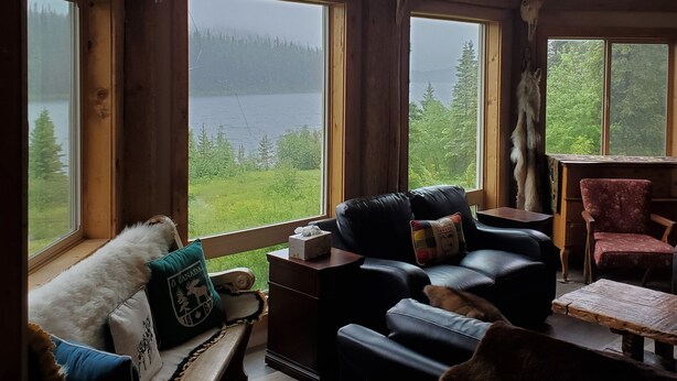 L’intérieur chaleureux d’un chalet, avec de grandes fenêtres, des poutres en bois, un canapé en cuir et une vue sur un lac et une forêt.