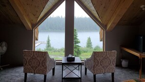 Une pièce avec un plafond haut et une grande fenêtre donnant sur un lac. On y trouve deux fauteuils à motifs et une petite table placée au centre.