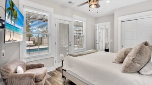 Une chambre à coucher comprenant un lit, une chaise, un ventilateur de plafond et de grandes fenêtres équipées de stores.