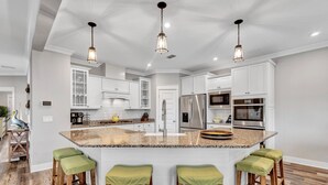 Une cuisine moderne avec des meubles blancs, un îlot en granit doté de tabourets, des appareils en acier inoxydable et des luminaires suspendus.