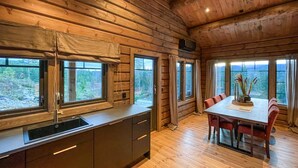 L’intérieur d’un chalet en bois comprend une cuisine et un coin repas, de grandes fenêtres offrant une vue sur un paysage boisé.