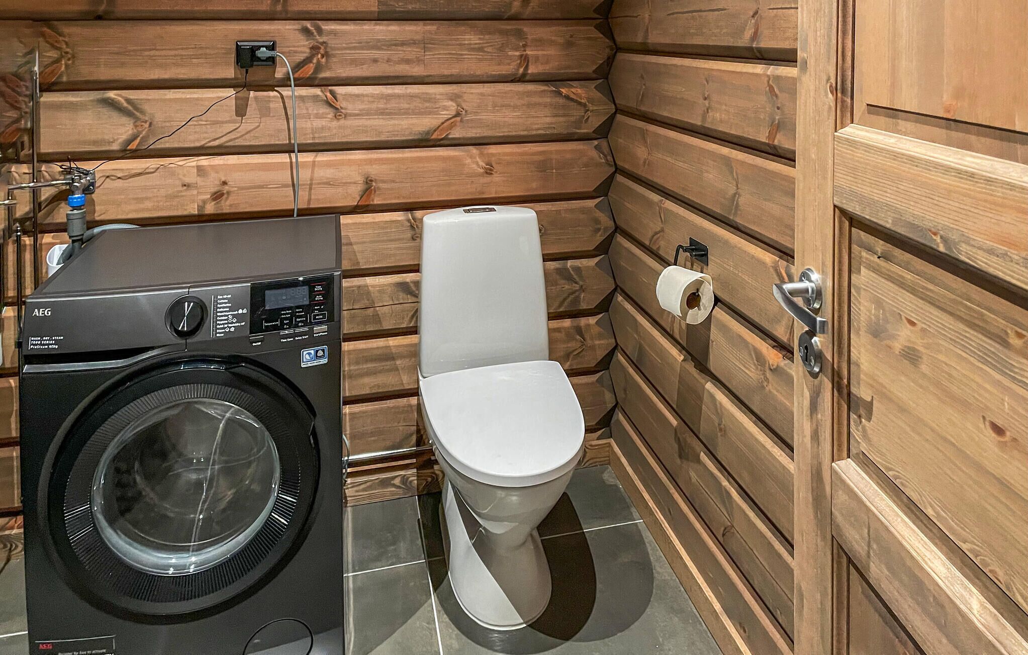 A compact bathroom with a washing machine, toilet, and wooden walls.