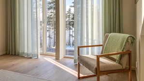 Ett rum med en trästol, stora fönster med tunna gardiner och utsikt över ett snötäckt landskap.