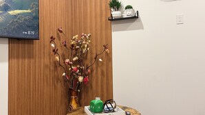 A room with a wooden panel wall, a mounted clock, a television displaying a scenic image, a vase with dried flowers, a small shelf with plants, and a table with board games. - 2-Bed, 2-Bath Home: Perfect for Work, Relaxation, and Exploring Calgary & Banff. (Calgary)
