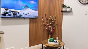 A living room with a flat-screen TV, a wooden accent wall, a clock, a small table with decorative items, and a brown leather sofa. - 2-Bed, 2-Bath Home: Perfect for Work, Relaxation, and Exploring Calgary & Banff. (Calgary)