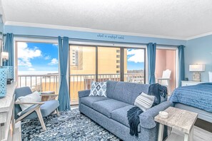 A room with a balcony, a sofa, a small table, a chair, and a bed.