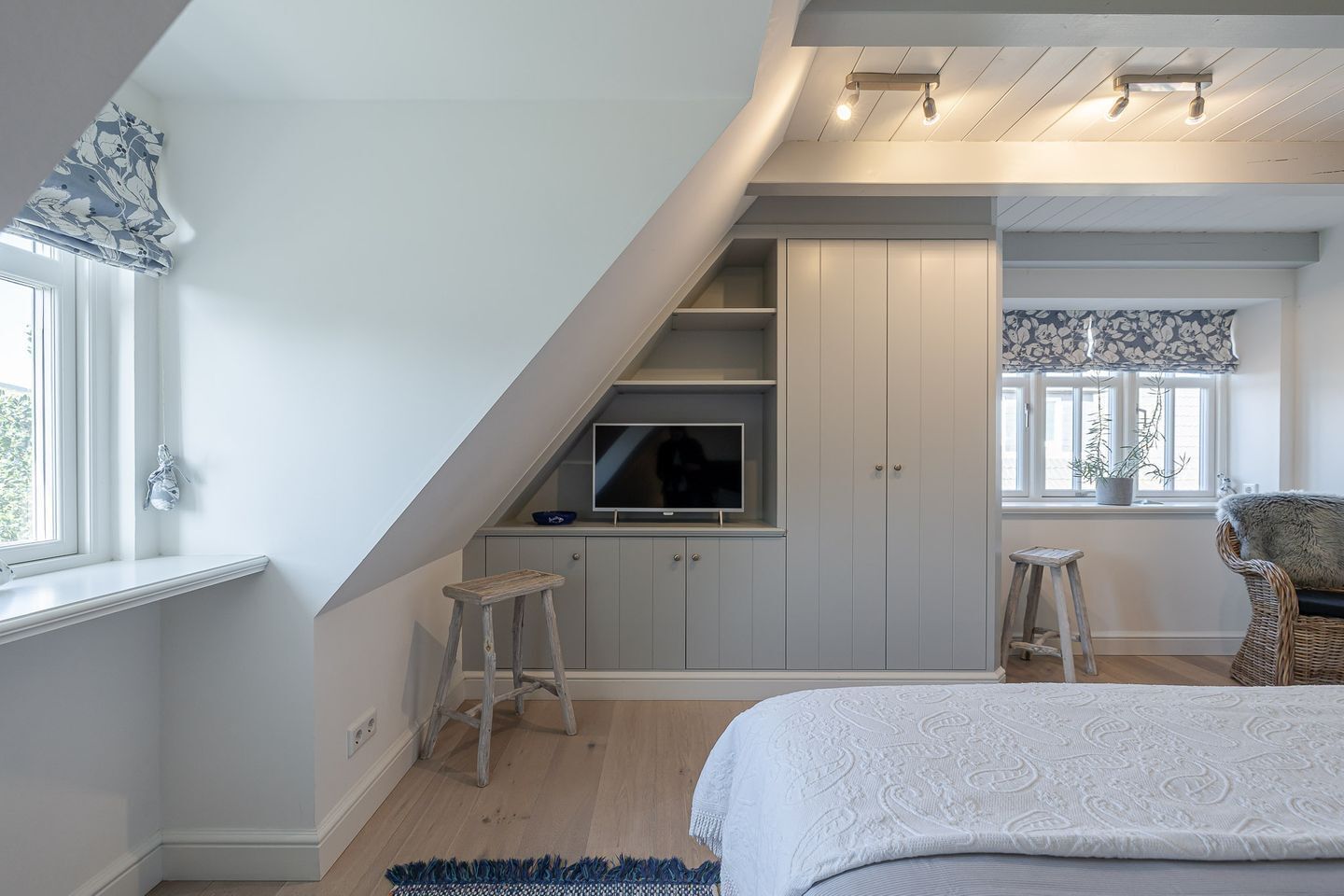 A bedroom with a sloped ceiling, a TV on a built-in shelf, a bed, a chair, and a window with a plant.